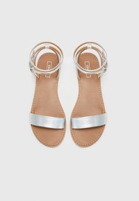 Zilveren platte sandalen met een brede metalen band en een enkelbandje. De zool is tan met zichtbare stiksels.