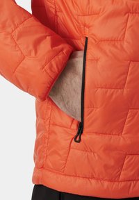 Chaqueta acolchada naranja con bolsillo de cremallera diagonal y textura acolchada. El material es suave, con un acabado ligeramente brillante.