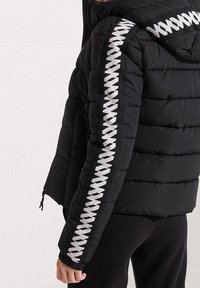 Chaqueta negra acolchada con capucha, que presenta un patrón de zigzag blanco texturizado en las mangas y un cierre frontal con cremallera.