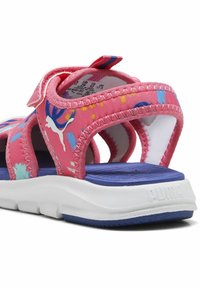 Rožiniai sportiniai sandalai su raštuotais dirželiais, turintys tvirtą Velcro užsegimą ir mėlyną tekstūruotą padą. Baltas padas su Puma logotipu.