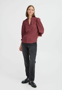 Bordeauxfarbene Bluse mit V-Ausschnitt, strukturierter Oberfläche und gerafften Puffärmeln. Trägt sie mit schwarzen Jeans und Loafers und steht vor einem weißen Hintergrund.