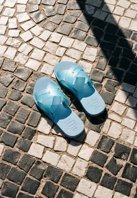 Sandales en caoutchouc bleu clair posées sur un pavement en pierre à motifs noir et blanc sous la lumière du soleil projetant une ombre partielle.