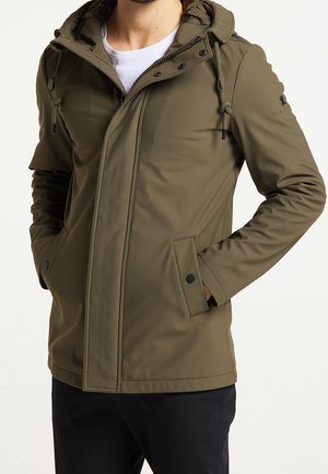 Hombre que lleva una chaqueta con capucha de color verde oliva con cremallera frontal, solapa con botones y manos en los bolsillos, combinada con pantalones negros y camisa blanca.