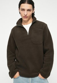 Suéter de forro polar marrón con cuello de media cremallera, que cuenta con un bolsillo frontal y tejido texturizado. El modelo lleva una camiseta blanca debajo.