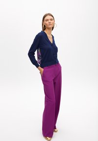 Navy button-up cardigan with v-neck and floral-patterned side panel, paired with high-waisted, wide-leg magenta trousers and metallic gold shoes.