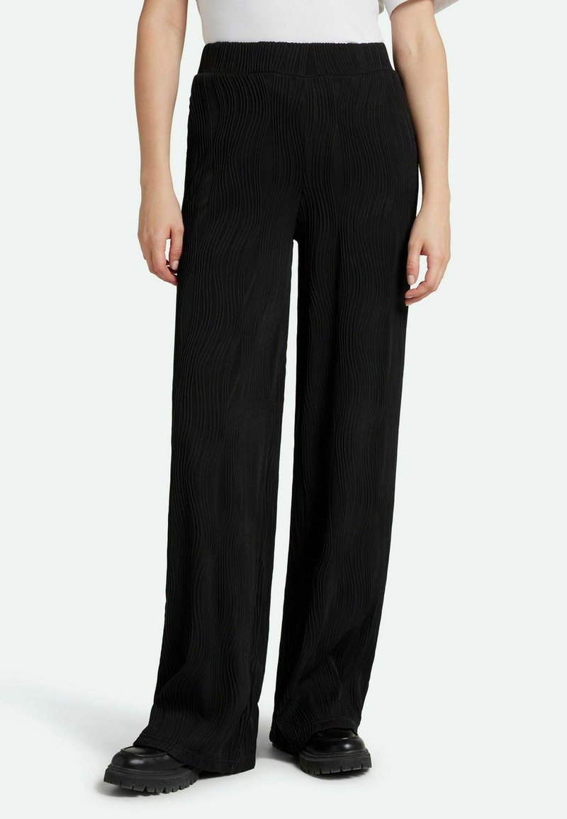 Black wide-leg pants with a textured ripple pattern and elastic waistband, paired with black platform shoes. Smooth fabric, casual design.
