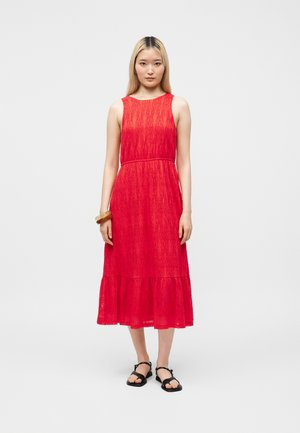 Naine seisab, kandes varrukateta punast tekstuuriga midi-kleit volangilise äärega, musti sandaale ja pruuni käeketti paremal randmel.