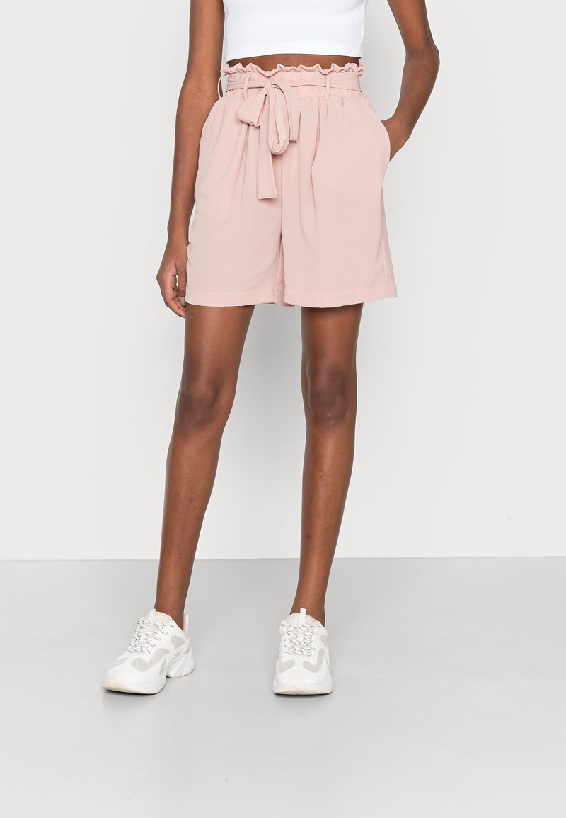 Personne portant un short taille haute rose pâle avec une ceinture nouée, associé à des baskets blanches et un haut blanc rentré.