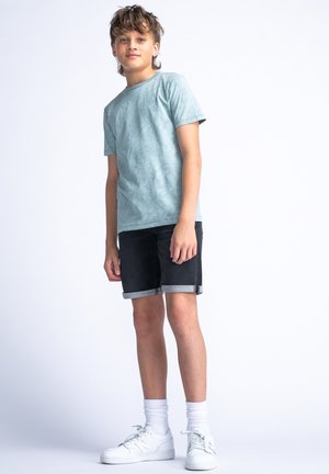 T-shirt texturé en teal, short en denim noir avec ourlet roulé, baskets blanches et chaussettes grises jusqu'à la cheville. Fond uni, pose debout.