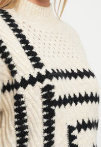 Cremefarvet striktrøje med høj hals, prydet med sorte zigzag- og geometriske mønstre samt en tekstureret overflade.