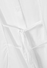 Hvidt stof med en glat tekstur har en bindelukning og draperinger i siderne. Sammensætningen er let og luftig, ideel til lag-på-lag brug.