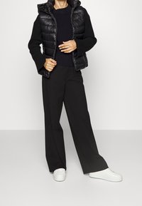 Veste noire matelassée avec capuche, associée à un pull côtelé bleu marine et un pantalon noir à jambes larges. Des baskets blanches complètent la tenue.