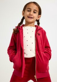 Rød velour hættetrøje med lynlås og et hjerteformet mærke, kombineret med en hvid skjorte med små røde hjerter samt matchende røde bukser. Glat tekstur.