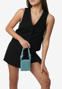 Piccola borsa rettangolare in materiale turchese scintillante, con manico a nodo torciglione e chiusura con zip, indossata con un gilet e una gonna neri.