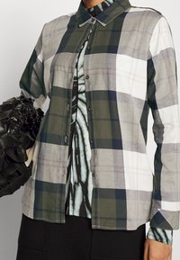 Koszula zapinana na guziki w kratę w kolorach zielonym, białym i szarym. Wykonana z tkaniny o gładkiej fakturze, zawiera kołnierz i guziki.