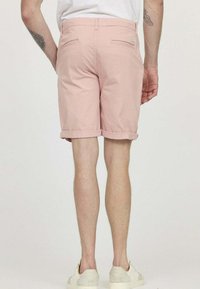 Shorts en coton rose avec un ourlet retroussé, comportant deux poches arrières et une petite étiquette de marque sur la ceinture. Portés avec des chaussures claires.