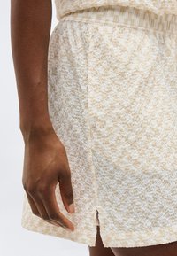 Χέρι σκουρόχρωμου δέρματος που ακουμπάει δίπλα σε μπεζ και λευκή υφασμάτινη φούστα με μικρή μπροστινή σχισμή και ελαστική μέση.