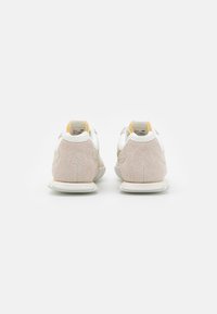 White and beige sneakers with a suede texture, featuring a padded collar, curved design, and a contrasting yellow accent on the heel.