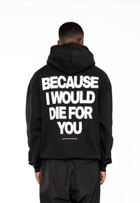 Sudadera negra con texto blanco en la parte de atrás que dice "PORQUE MORIRÍA POR TI", acentuada por un pequeño logo dorado en la parte inferior.
