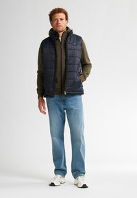 Navy Weste über einem grünen Kapuzenpullover, kombiniert mit hellblauen Jeans und weißen Turnschuhen. Die Weste hat einen Reißverschluss und Steppnähte.