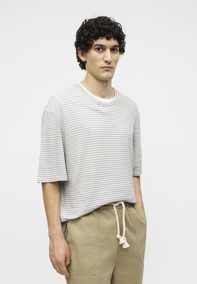 Νέος άνδρας με σγουρά μαλλιά, που φορά ένα ανοιχτό γκρι και λευκό ριγέ πουκάμισο και μπεζ παντελόνι με ζώνη από σκοινί, στέκεται με το ένα χέρι στην τσέπη.