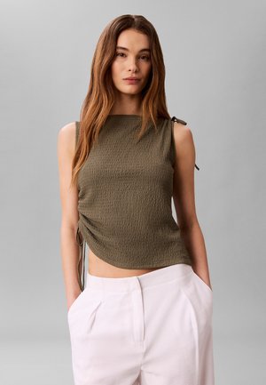 Vrouw met lang bruin haar die een mouwloos olijfgroen textuur topje draagt en witte broek met hoge taille met handen in de zakken.