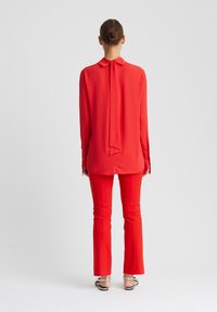Rote Bluse mit einer Schleife am Kragen, kombiniert mit roten ausgestellten Hosen. Glatter Stoff, lange Ärmel und Knopfdetails an den Manschetten.