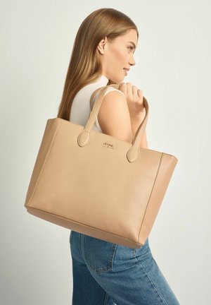 Kobieta z długimi włosami, ubrana w bezrękawnik i dżinsy, niosąca na ramieniu dużą beżową skórzaną torbę typu tote.