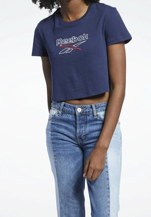 Woman wearing navy blue cropped Reebok T-shirt and high-waisted two-tone denim jeans, standing against a plain white background.