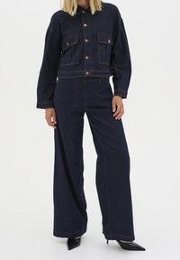 Nainen, jolla on tummansininen farkkutakki ja leveälahkeiset farkut, yhdistettynä mustiin teräväkärkisiin korkokengiin, seisoo valkoista taustaa vasten.