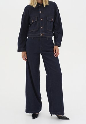 Mujer vestida con una chaqueta de denim azul oscuro y jeans de pierna ancha, combinados con zapatos de tacón alto negros de punta afilada, de pie contra un fondo blanco.