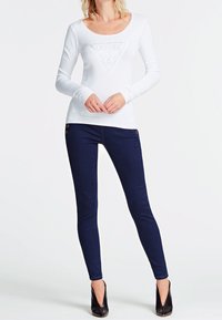Frau in einem weißen langärmeligen Oberteil mit einem dreieckigen Logo und dunklen Skinny-Jeans, die mit verschränkten Händen und schwarzen Absätzen steht.