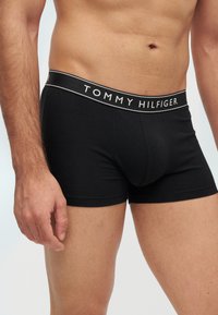 Άνδρας κορμός που φοράει μαύρα εσώρουχα Tommy Hilfiger με ορατό σκουλαρίκι στον αφαλό, σε απλό φωτεινό φόντο.