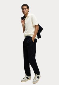 Hvid ribbet polo, marineblå bukser og træningssko med sorte accenter. Modellen holder en jakke, hvilket viser et afslappet, moderne outfit.