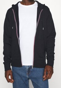 Sudadera negra con cremallera hecha de un tejido suave, con un bolsillo canguro dividido, que presenta un acento colorido a lo largo de la cremallera. Llevada sobre una camiseta blanca.