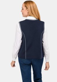 Vrouw met lang rood haar, gekleed in een marineblauwe mouwloze vest over een witte blouse met lange mouwen en blauwe spijkerbroeken, met haar rug naar de camera gekeerd.