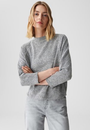 Grauer strukturierter Pullover mit hohem Kragen, langen Ärmeln und einem lockeren Schnitt, kombiniert mit hellgrauen Jeans, vor einem einfarbigen Hintergrund.