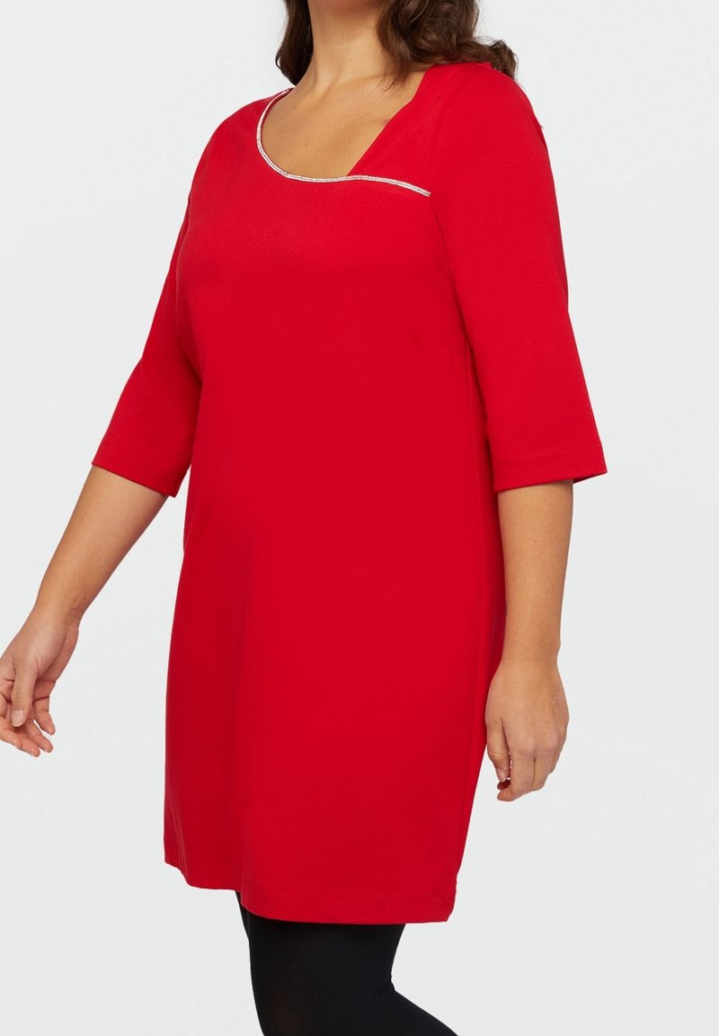 Femme portant une robe rouge longueur genoux avec des manches trois-quarts et un col arrondi avec une bordure blanche, debout devant un fond uni.