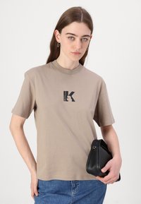 Bežna bombažna majica s kratkimi rokavi in okroglim izrezom, ki prikazuje črno grafiko z obliko "K". Nosi se z modrimi kavbojkami in črno torbico.