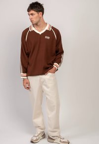 Brązowa prążkowana bluzka z długim rękawem, z białym i pomarańczowym paskowanym kołnierzem i mankietami, zestawiona z luźnymi jasnymi spodniami i trampkami.