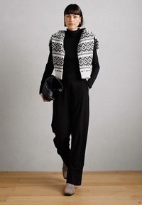 Pulover negru cu gât sub o vestă cropt cu model gri și alb, asortat cu pantaloni largi negri și cizme gri până la gleznă. Ținând o geantă neagră.