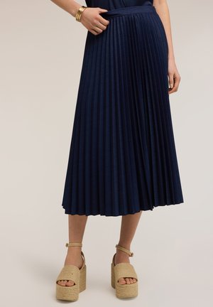 Kobieta ubrana w granatową plisowaną spódnicę midi, beżowe sandały na platformie oraz złote bransoletki na nadgarstku, stojąca na tle jednolitego tła.