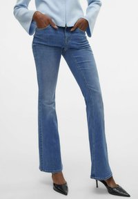 Modre flare jeans iz denima z nizkim pasom, sprednjimi žepi in gladko teksturo. Kombinirane z črnimi čevlji s koničastim prstom.