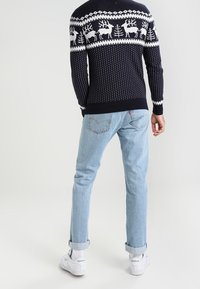 Navy gebreide trui met witte rendieren en boompatronen, gecombineerd met lichtblauwe spijkerbroeken en witte sneakers met groene accenten.