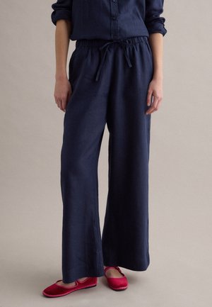 Persona con pantaloni larghi blu navy con fascia elastica e coulisse, abbinati a scarpe basse rosa acceso su sfondo semplice.