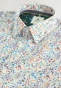Chemise blanche à boutons avec un imprimé floral composé de fleurs et de feuilles multicolores. Le col présente une doublure intérieure fleurie contrastante de couleur verte.