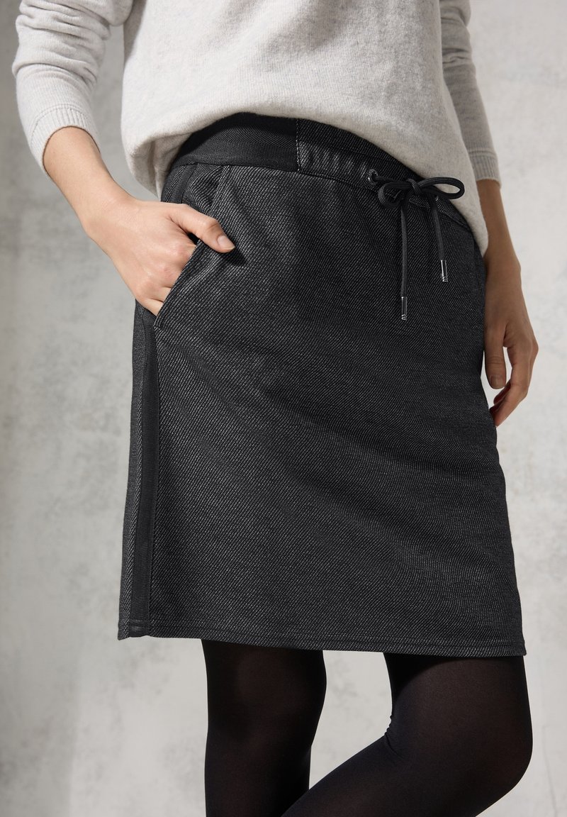 Personne portant une jupe gris foncé mi-longue avec taille à cordon de serrage et poches latérales, associée à des collants noirs et un pull gris clair.