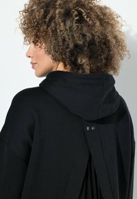Černá mikina s volným střihem, s detailem kapuce a rozparkem vzadu. Obsahuje knoflíky a skladby pro přidání textury a designového zájmu.