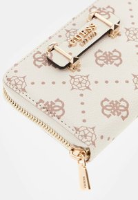 Portafoglio bianco Guess con motivo logo beige, zip dorata e placca, primo piano che mostra la texture e i dettagli metallici con il marchio.