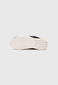 Zwarte sneaker met een gestructureerde bovenkant, voorzien van een witte rubberen zool met een patroon. Merken zichtbaar op de zool.